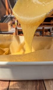 Yellow cake batter is being poured from a bowl into a parchment paper-lined baking dish on a wooden surface, with a warm kitchen setting in the background.