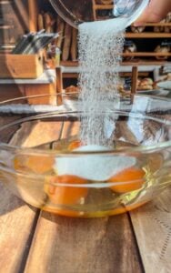 A close-up of sugar being poured from a small glass container into a clear bowl containing three raw eggs on a wooden surface, with a bakery-style kitchen in the background.