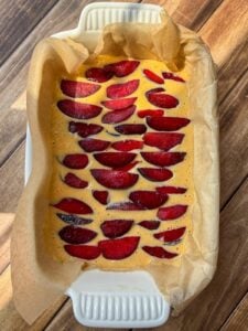 A rectangular baking dish lined with parchment paper holds an unbaked batter topped with neatly arranged slices of red plums, ready to be baked, on a wooden surface.