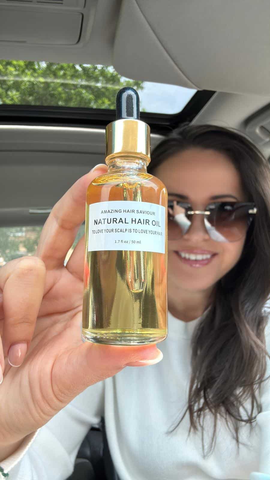 A woman with dark hair and sunglasses holds up a bottle of "Natural Hair Oil" with a dropper lid inside a car, smiling as she displays the product toward the camera.