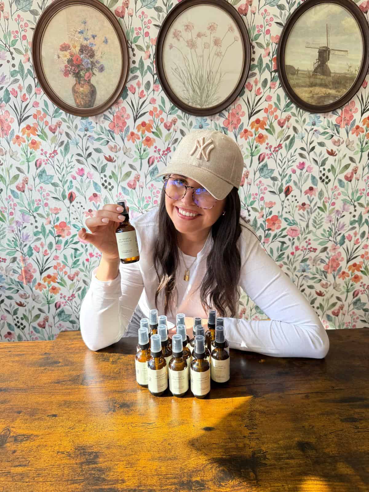 A woman in a beige cap and glasses sits at a wooden table with several small bottles in front of her, holding one up and smiling. The background features floral wallpaper and framed art.