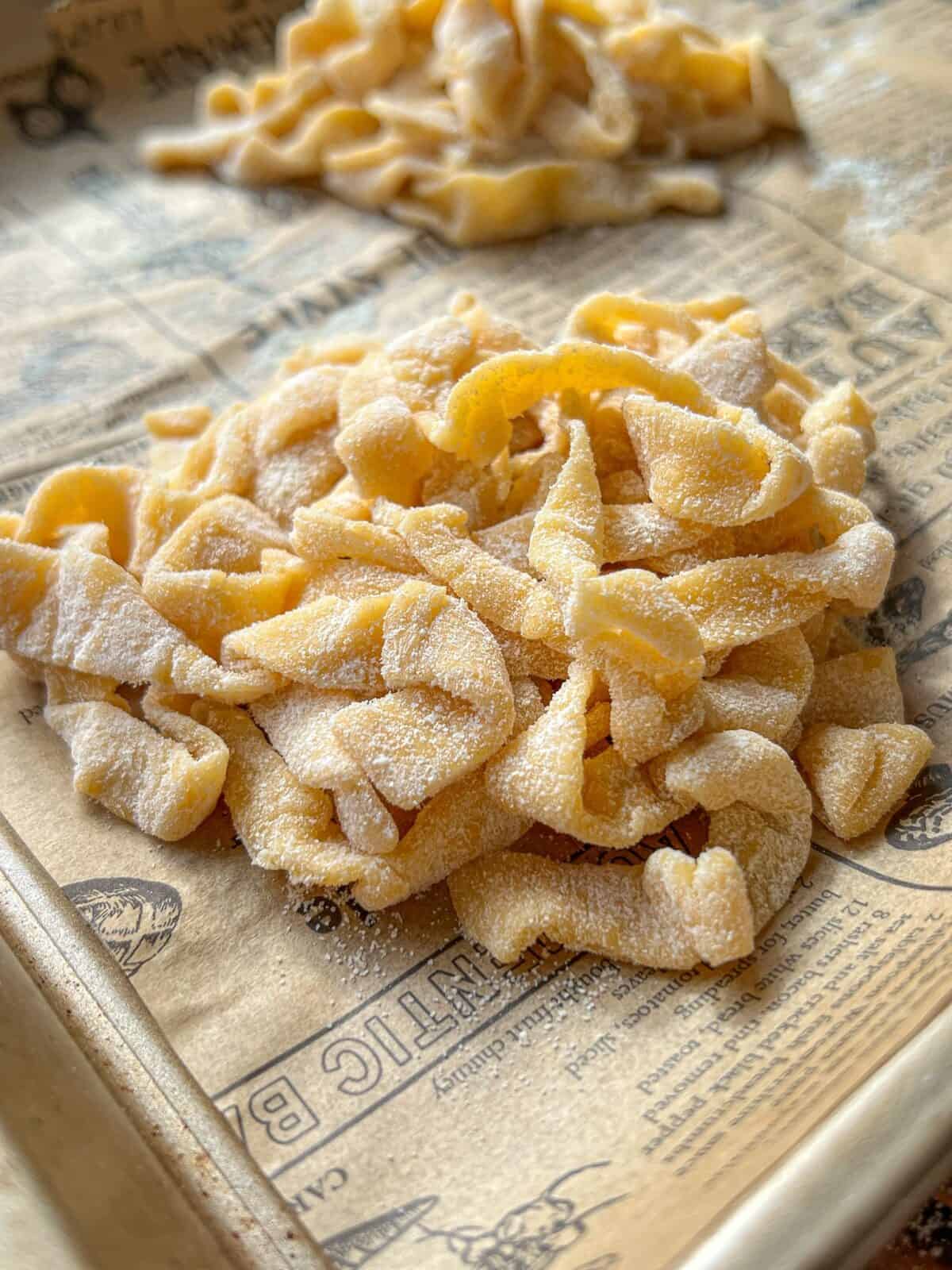 A close-up of fresh, uncooked homemade pasta dusted with flour, resting on parchment paper printed with vintage-style illustrations and text.