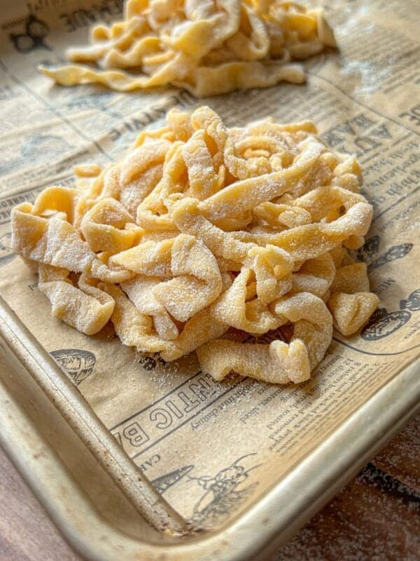 Freshly made, uncooked pasta noodles dusted with flour are piled on a baking sheet lined with vintage-style paper. More pasta is visible in the background.