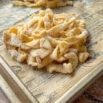 A close-up of a pile of uncooked, homemade pasta noodles dusted with flour on a baking sheet lined with printed parchment paper. Another small pile of noodles is in the background.