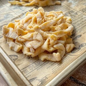 A close-up of a pile of uncooked, homemade pasta noodles dusted with flour on a baking sheet lined with printed parchment paper. Another small pile of noodles is in the background.