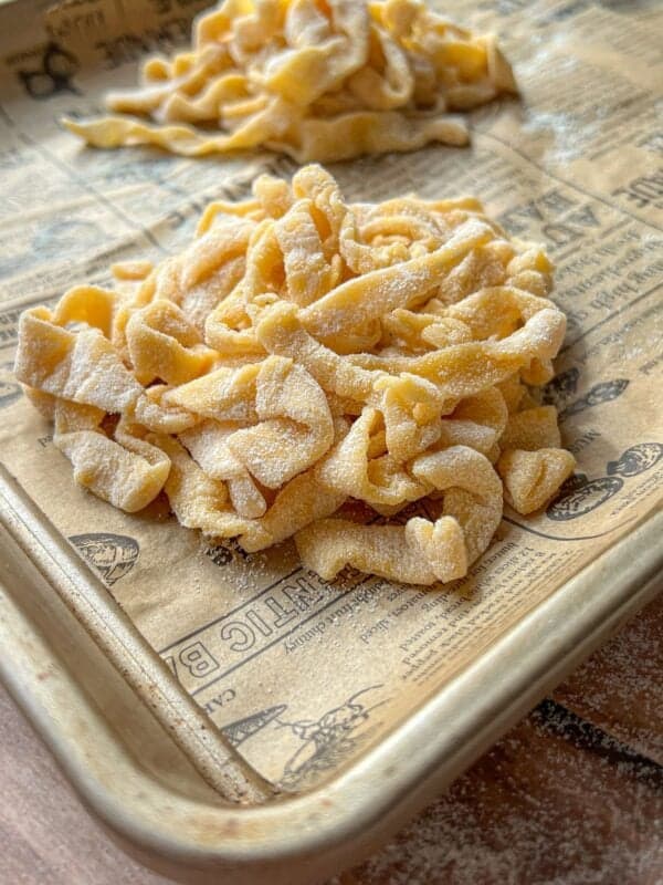 A close-up of a pile of uncooked, homemade pasta noodles dusted with flour on a baking sheet lined with printed parchment paper. Another small pile of noodles is in the background.