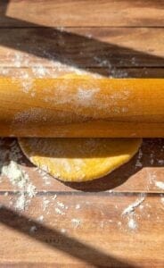 A wooden rolling pin flattens a ball of dough on a floured wooden surface, with sunlight casting shadows across the workspace.