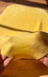 Close-up of hands stretching a thin, yellow sheet of fresh pasta dough, with a larger piece of pasta dough resting on a wooden surface in the background. Sunlight highlights the dough’s smooth texture.