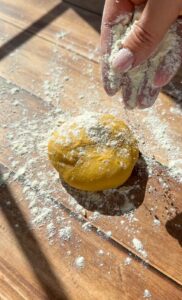 A hand sprinkles flour over a ball of yellow dough on a wooden surface, with scattered flour around, in bright natural sunlight.