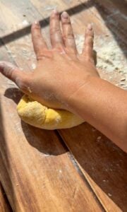 A hand is kneading dough on a floured wooden surface in bright sunlight, with flour scattered around.