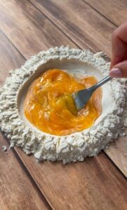 A hand uses a fork to whisk eggs in the center of a mound of flour on a wooden surface, preparing dough.
