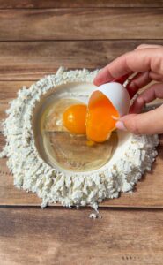 A hand cracks an egg into a well of flour on a wooden surface. The flour forms a circle with the egg yolk and whites in the center, ready for mixing, suggesting the start of a cooking or baking process.
