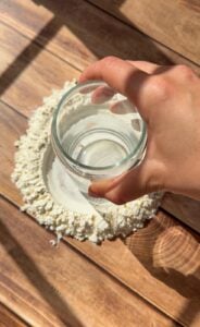 A hand holds a glass of water above a mound of flour with a well in the center, placed on a wooden surface in sunlight.