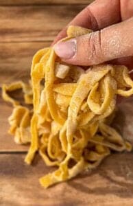 A hand holding freshly made, uncooked fettuccine pasta on a wooden surface, with flour visible on the fingers and pasta.