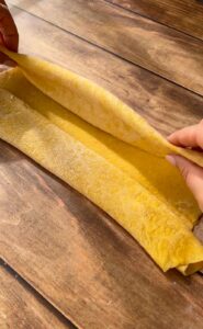 Hands carefully lift and fold a sheet of freshly rolled yellow pasta dough on a wooden surface, preparing it for cutting or shaping.