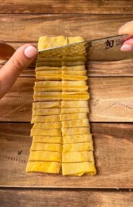 A person slices a stack of thin yellow dough sheets into strips with a knife on a wooden surface, preparing homemade pasta or noodles.