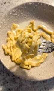 A close-up of homemade pasta noodles in a creamy yellow sauce on a speckled ceramic plate, with a fork lifting a portion above the dish.