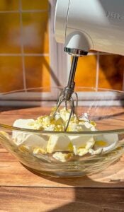 A hand mixer with metal beaters is positioned above a glass bowl containing cream cheese and butter, ready to be blended. Warm sunlight streams through a window in the background.