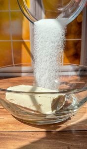 Granulated sugar is being poured from a glass bowl onto a block of cream cheese in a clear glass mixing bowl, set on a wooden surface with a sunny window in the background.