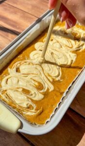 A hand swirls cream cheese into pumpkin batter with a skewer in a loaf pan, creating a marbled effect before baking. The pan sits on a wooden surface.