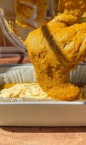 Thick, golden-orange batter is being poured from a bowl into a parchment-lined baking dish, ready to be baked. The texture is airy and slightly bubbly, with sunlight highlighting the rich color.