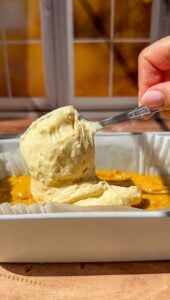 A hand holds a spoonful of thick, creamy batter over a rectangular baking dish with a yellow mixture inside, set on a wooden surface near a sunlit window.