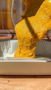 Bright yellow-orange batter is being poured from a bowl into a rectangular paper-lined baking dish, with sunlight casting shadows across the scene.