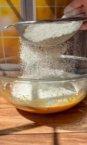 A close-up of a hand sifting flour through a metal sieve into a glass bowl containing a yellow batter, with sunlight streaming through a window and yellow tiles in the background.