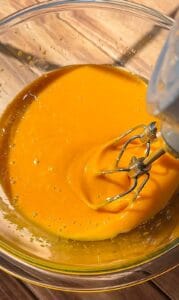 A close-up of an electric mixer blending a thick, bright orange batter in a clear glass bowl placed on a wooden surface.