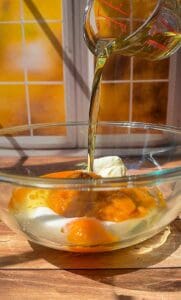 A glass measuring cup pours oil into a glass bowl containing eggs, sugar, and a dollop of cream, with sunlight shining through a window in the background.