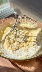A hand mixer blending flour into a creamy batter in a glass bowl on a wooden surface, with sunlight streaming in from a window nearby.