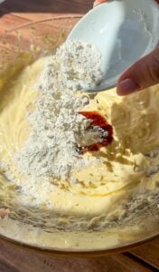 A hand pours flour from a small white bowl into a glass mixing bowl filled with creamy batter, ready to be mixed together.