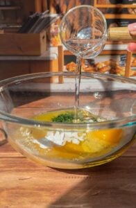 A hand pours water from a small glass cup into a large glass bowl containing eggs, flour, and herbs on a wooden countertop, preparing the base for Authentic Matzo Ball Soup in a cozy bakery setting.
