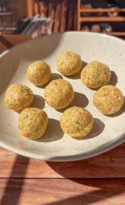A beige plate holds eight round, yellowish snack balls with green flecks, arranged neatly—reminiscent of Authentic Matzo Ball Soup. The plate sits on a wooden surface, set against a softly blurred, rustic background.