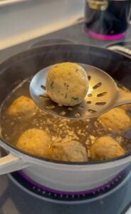 A slotted spoon holds a matzo ball over a pot of simmering broth, with more matzo balls cooking in the liquid below on a stovetop. Steam rises from the pot, capturing the warmth of authentic matzo ball soup, hot and freshly cooked.