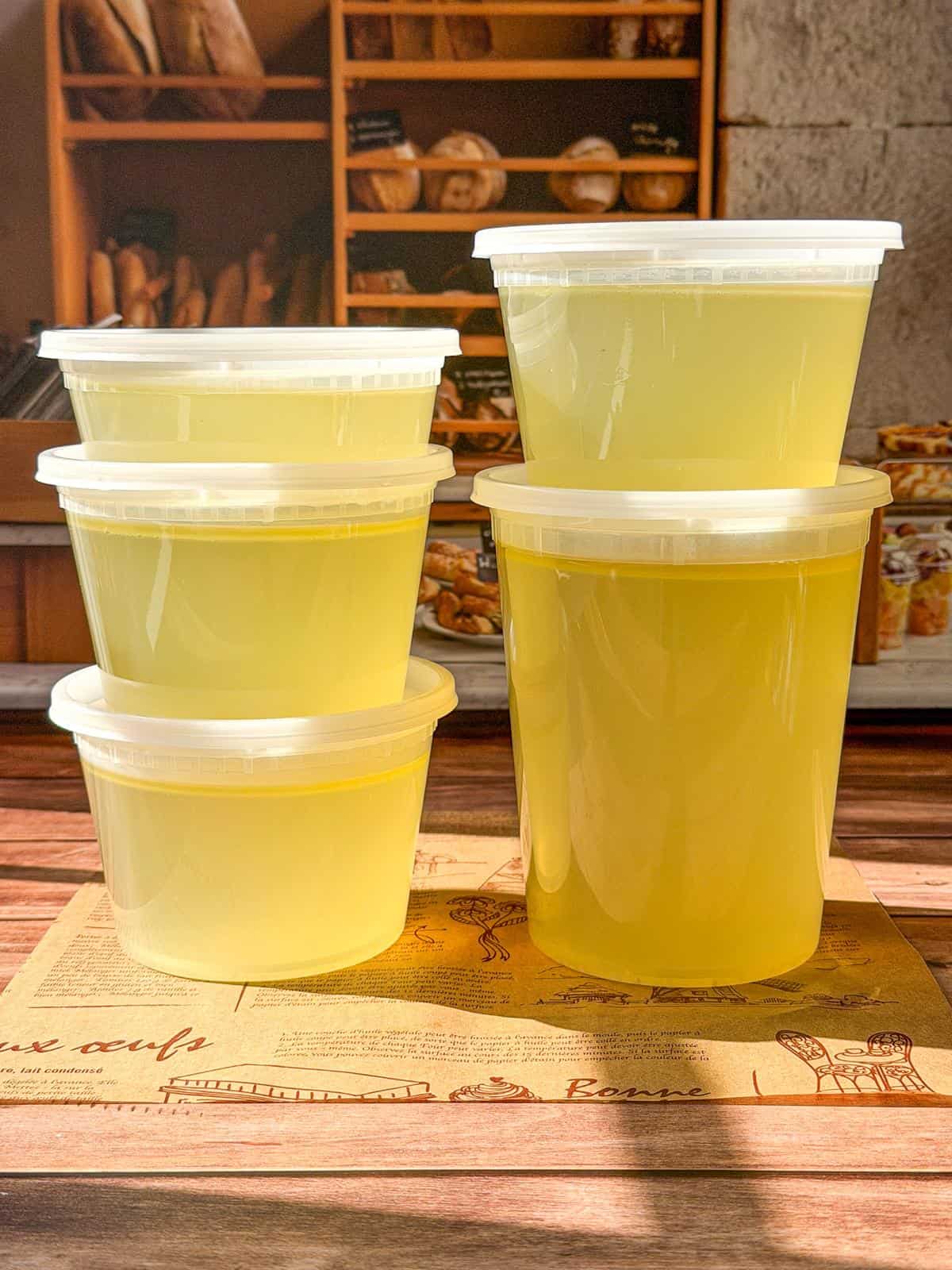 Five plastic containers filled with yellow liquid labeled Easy Homemade Chicken Broth sit stacked on a table. In the bakery background, shelves of bread and pastries glow in the sunlight, creating a warm and inviting atmosphere.