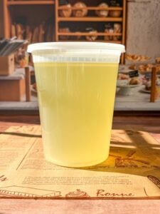 A clear plastic container filled with light yellow Easy Homemade Chicken Broth sits on a table, with a bakery display and baked goods visible in the blurred background.