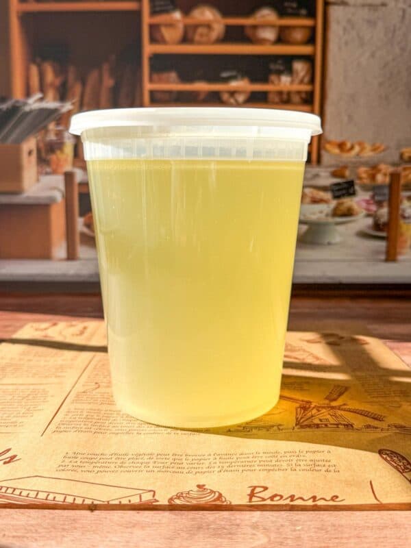 A clear plastic container filled with light yellow Easy Homemade Chicken Broth sits on a table, with a bakery display and baked goods visible in the blurred background.