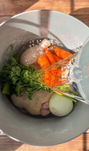 A pot contains raw whole chicken, carrots, an onion, and fresh herbs as water is poured in from a pitcher—perfect for starting an easy homemade chicken broth.