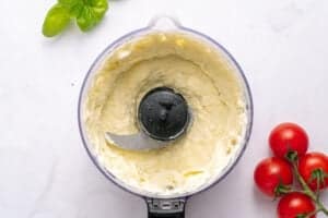 A food processor bowl filled with creamy ricotta cheese mixture, perfect for your next Tomato Sandwich Recipe, sits beside cherry tomatoes and fresh basil leaves on a white surface.