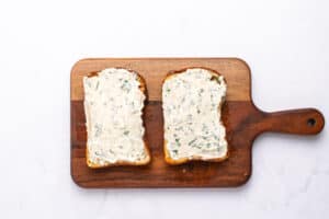 Two slices of toasted bread with a creamy white herb spread sit side by side on a wooden cutting board—a perfect start to any Tomato Sandwich Recipe—set against a clean white background.