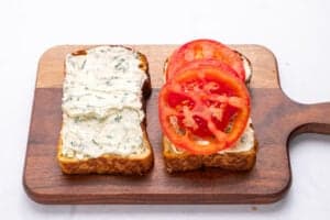 Two slices of bread on a wooden cutting board, one spread with herb cream cheese and the other topped with cream cheese and sliced tomatoes—perfect for trying out your next Tomato Sandwich Recipe.