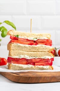 Two halves of a tomato sandwich with white bread, mayonnaise, and herbs, stacked on top of each other with a wooden skewer, on a wooden board. Fresh tomatoes and basil are in the background.
