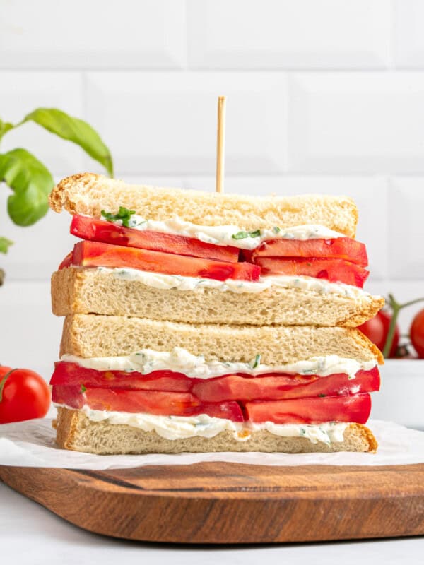 Two halves of a tomato sandwich with white bread, mayonnaise, and herbs, stacked on top of each other with a wooden skewer, on a wooden board. Fresh tomatoes and basil are in the background.