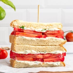A stacked tomato sandwich with thick tomato slices, creamy spread, and herbs on white bread, held together with a skewer, sits on parchment paper atop a wooden board. Try this classic Tomato Sandwich Recipe—fresh tomatoes and basil complete the scene.