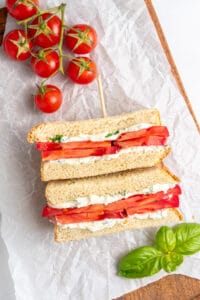 A Tomato Sandwich Recipe features thick tomato slices and a creamy filling, cut in half and stacked on parchment paper. Fresh basil leaves and a cluster of ripe tomatoes rest nearby on a wooden board.
