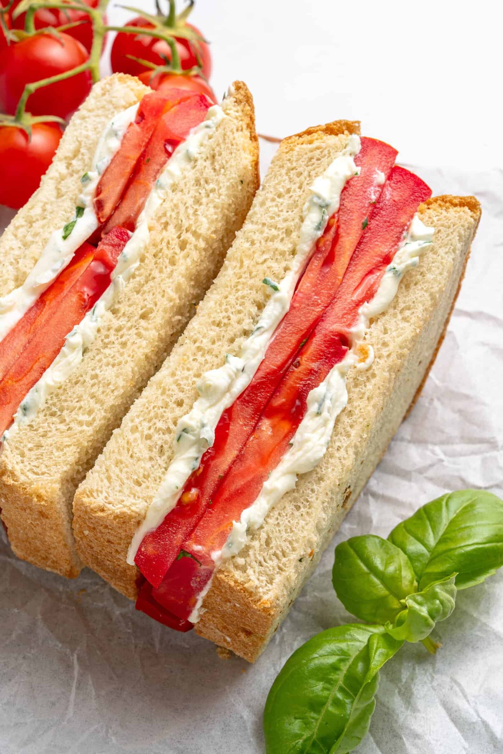 Two thick slices of sandwich bread filled with layers of fresh tomato and herbed cream cheese are placed on parchment paper, with vine tomatoes and fresh basil nearby.