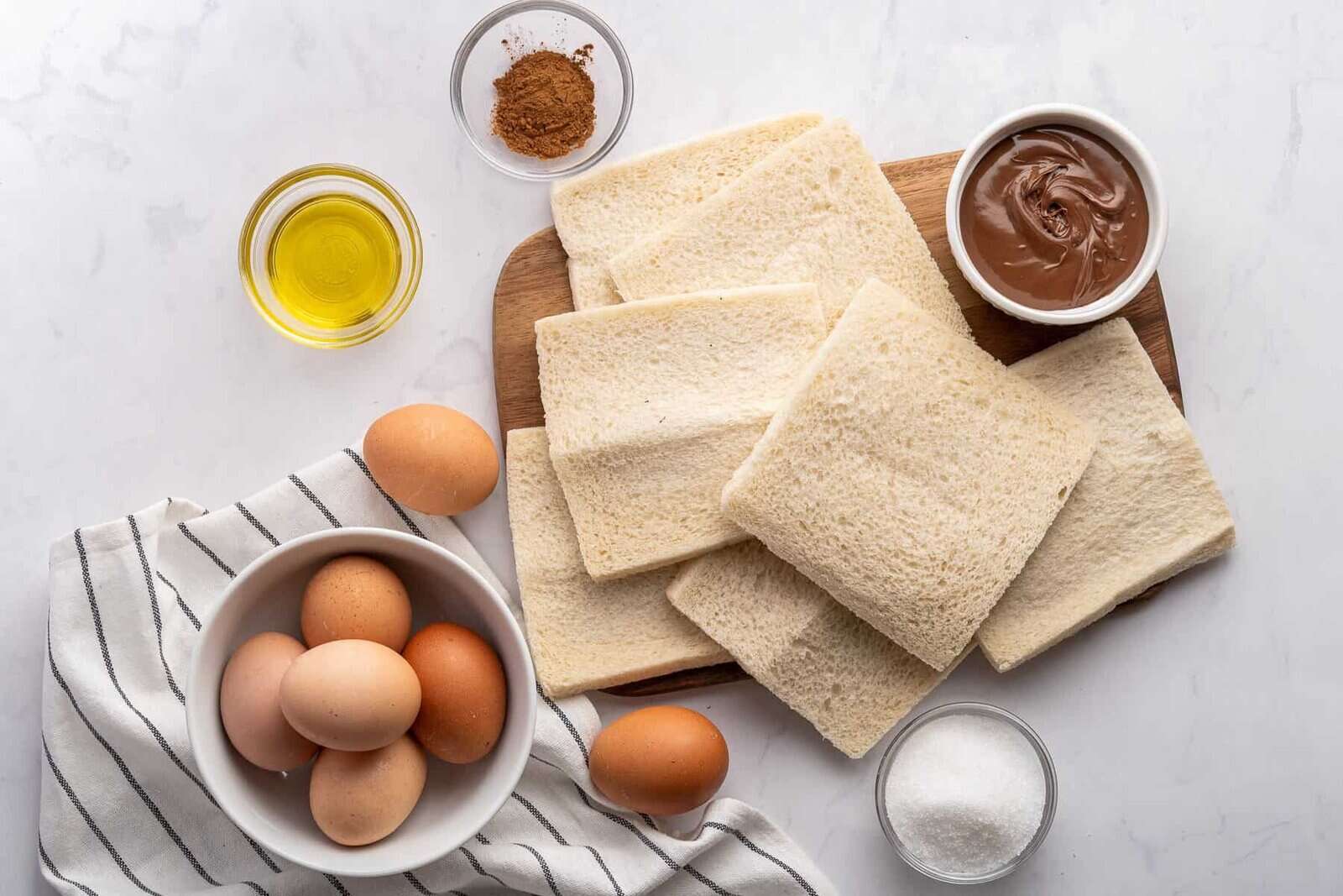 Several slices of white bread on a cutting board, surrounded by a bowl of eggs, a small bowl of chocolate spread, a bowl of oil, a dish with cinnamon, and a bowl of sugar on a white surface with a striped cloth.