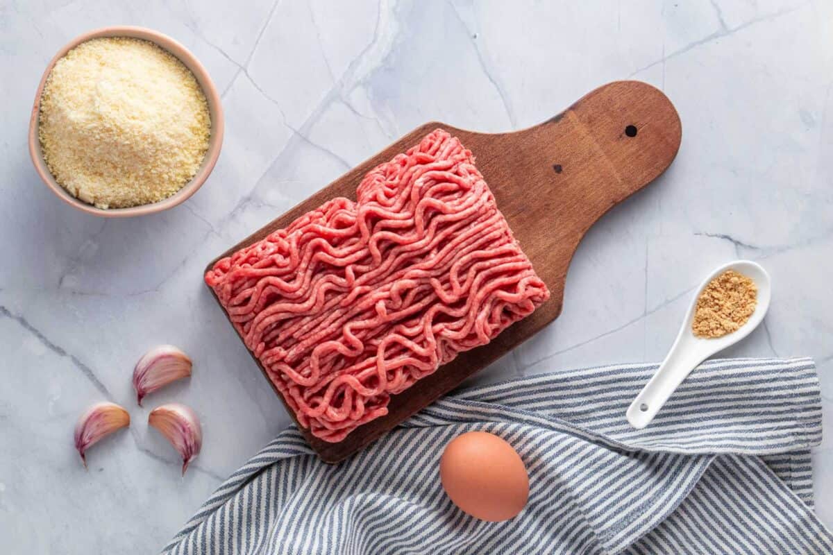 Raw ground beef on a wooden cutting board, a bowl of grated cheese, three garlic cloves, an egg, a spoonful of breadcrumbs, and a striped towel on a marble surface.