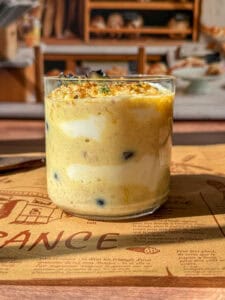 A glass filled with layers of creamy yellow and white yogurt, some blueberries, and a dusting of spices on top, placed on a brown placemat with bakery shelves visible in the blurred background.